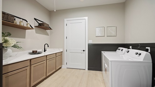 Large laundry room with a sink, light stained cabinets, white and gray quartz countertops, upgraded washer and dryer, white walls, and light vinyl wood flooring.