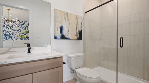 Secondary bathroom with a sink, white and gray quartz countertops, walk-in shower with a glass door, light stained cabinets, white walls, and light vinyl wood flooring.