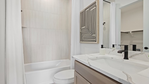 Secondary bathroom with a sink, white and gray quartz countertops, shower and tub combo, light stained cabinets, white walls, and light vinyl wood flooring.