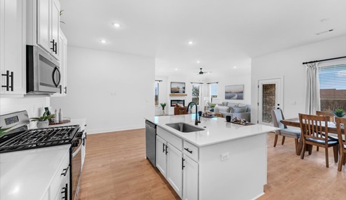 Modern kitchen with white cabinets, stainless steel appliances, and a sleek white quartz countertop. Features include a center island with a built-in sink, a gas stove, and a decorative clock on the wall. The space is accented with plants and minimalistic decor.