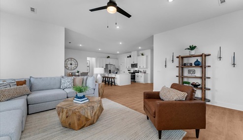 Spacious open-concept living area with a light gray sectional sofa, a wooden geometric coffee table, and a leather accent chair. The room features modern decor, a ceiling fan, and a view of the adjacent kitchen with white cabinetry and a center island. A wooden shelf and wall-mounted candle holders add a decorative touch.