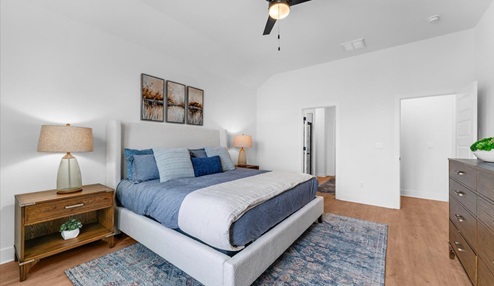 Bright and airy bedroom with a large upholstered bed featuring blue and neutral bedding, flanked by wooden nightstands with matching lamps. The room includes a vaulted ceiling with a ceiling fan, large windows with blue curtains, and a decorative rug. A potted plant and a wooden dresser add warmth and natural elements.