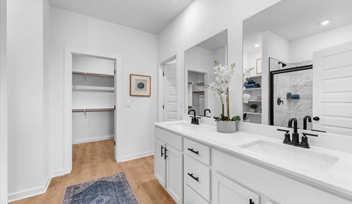 Modern bathroom with a double vanity featuring white countertops and black faucets, large mirrors, and decorative orchid accents. A glass-enclosed shower is visible, and the space connects to a bedroom with large windows. Built-in shelving adds functionality, and a blue patterned rug completes the design.