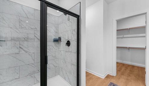 Modern bathroom with a double vanity featuring white countertops and black faucets, large mirrors, and decorative orchid accents. A glass-enclosed shower is visible, and the space connects to a bedroom with large windows. Built-in shelving adds functionality, and a blue patterned rug completes the design.