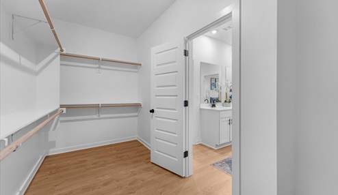 Spacious walk-in closet with white walls, wooden shelving, and hanging rods, featuring a view into a modern bathroom with double sinks and a vanity area. The closet has wood flooring and ample storage space.