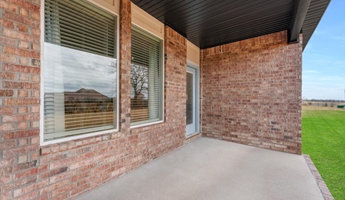 Covered outdoor patio with brick walls, large windows, and a concrete floor, overlooking a well-maintained green lawn and a scenic backyard view.