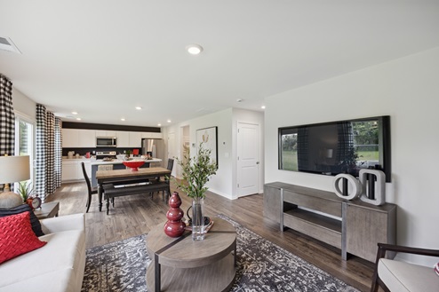 View of open concept living area from living room. Round brown coffee table and tv on wall over brown console table. Light brown dining table with dark brown bench and chairs. Kitchen with white cabinetry and stainless appliances against black accent wall. White walls, brown vinyl plank floors. Windows with white and black check patterned curtains along left wall.