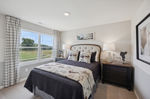 Bedroom with upholstered bed and purple bedding. Brown bedside tables with white lamps. Large window with black and white curtains on left wall. Beige walls and beige carpet.