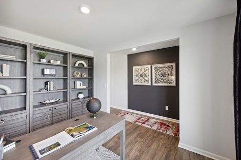 Home office with white walls and brown vinyl plank floors. Light brown desk across from grey wall shelving unit with decor. View of entry of home with black accent wall and black and white artwork over red and ivory runner rug.