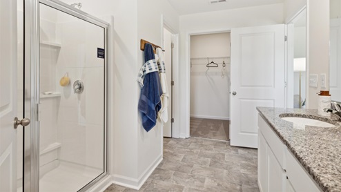 Primary bathroom with tile flooring, a double vanity with dark countertops upon white, shaker-style cabinetry and a walk-in shower with framed glass door.