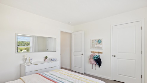Secondary bedroom with pastel striped bedspread, hooks on the wall for backpack and cheer bag, and a dresser boasting trophies beneath a large mirror.