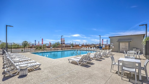 Community swimming pool at Tempo Trails featuring clear blue water, surrounded by lounge chairs and a secure fence. The pool area includes shaded seating, restroom facilities, and is set against a backdrop of modern residential buildings and clear blue skies.
