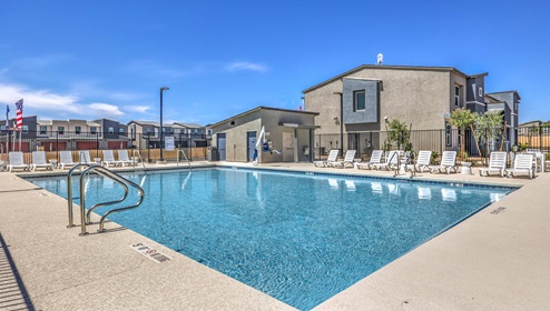 Community swimming pool at Tempo Trails featuring clear blue water, surrounded by lounge chairs and a secure fence. The pool area includes shaded seating, restroom facilities, and is set against a backdrop of modern residential buildings and clear blue skies.