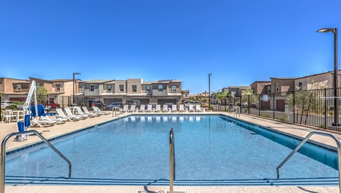 Community swimming pool at Tempo Trails featuring clear blue water, surrounded by lounge chairs and a secure fence. The pool area includes shaded seating, restroom facilities, and is set against a backdrop of modern residential buildings and clear blue skies.