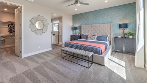 The main bedroom in our Heartland Manor 2754 Plan features plush carpet flooring, a spacious layout, and large windows that bring in natural light. A ceiling fan is centrally located for comfort, and an open doorway leads to the en-suite bathroom. The room also includes a walk-in closet, providing ample storage space.