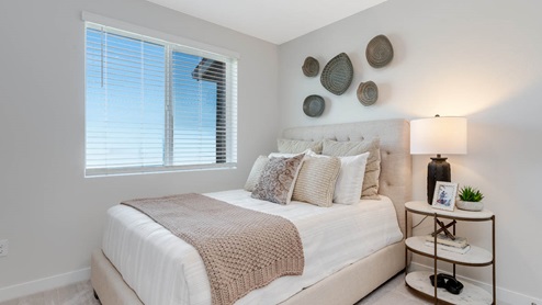 Scholls Meadow bedroom with end table and lamp, bed frame with grey light brown bed sheets and pillows, carpet, and windows