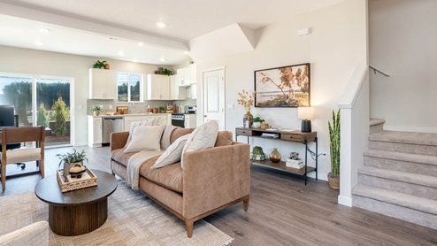 Scholls Meadow Great room with rug, coffee table, decorative middle piece, console table with lamp, plants and paintings, couch with throw blanket, laminate flooring, poster, lamp, window, and staircase, and view of kitchen