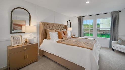 Primary bedroom with a large bed with upholstered headboard flanked by two nightstands with an accent chair in the corner next to the double window.