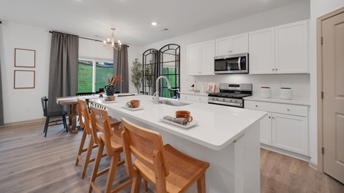 A spacious kitchen with white, shaker-style cabinets, quartz countertops, a large center island with undermount sink and seating for three, and a stainless-steel appliance package that flows into an adjoining dining area that boasts a wooden table surrounded by six chairs.