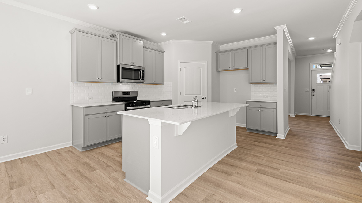 The kitchen has an island with a sink, granite countertops, grey shaker-style cabinets, a subway tile backsplash, stainless steel appliances, and a view of the front door with a transom.