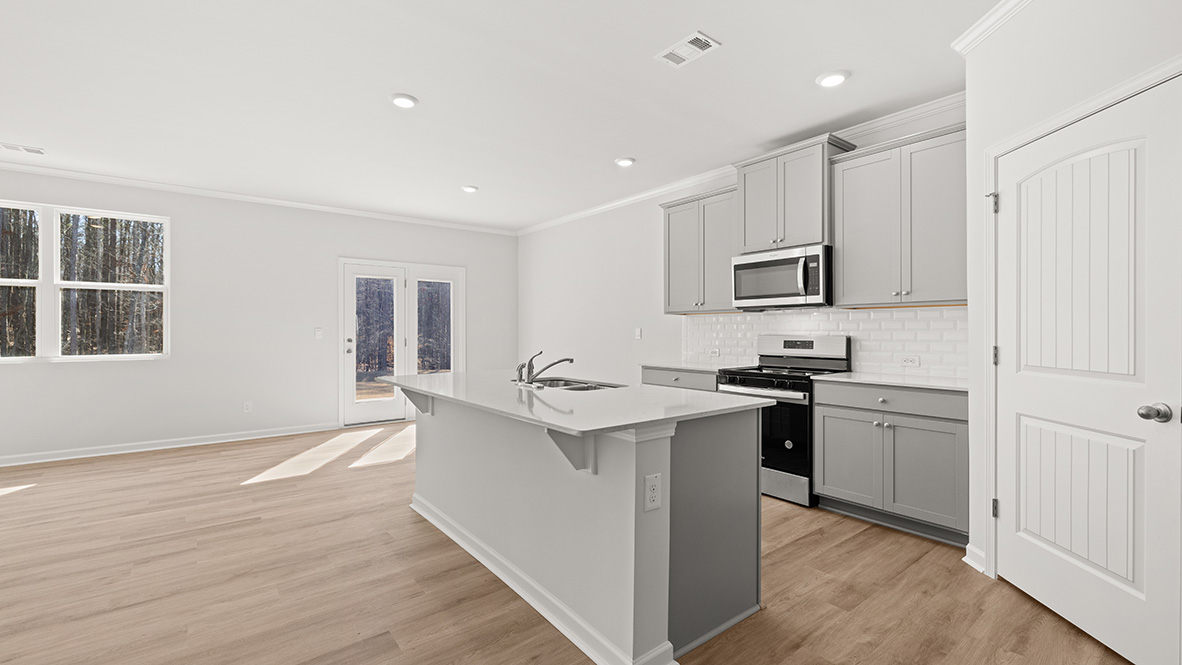 This is a view of the kitchen and dining room through a glass door. The kitchen has an island with a sink, granite countertops, grey shaker-style cabinets, a subway tile backsplash, and stainless steel appliances.