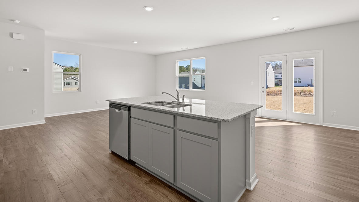 A bright, modern kitchen features a large island with a sink and dishwasher. Sunlit hardwood floors complement white walls and large windows.