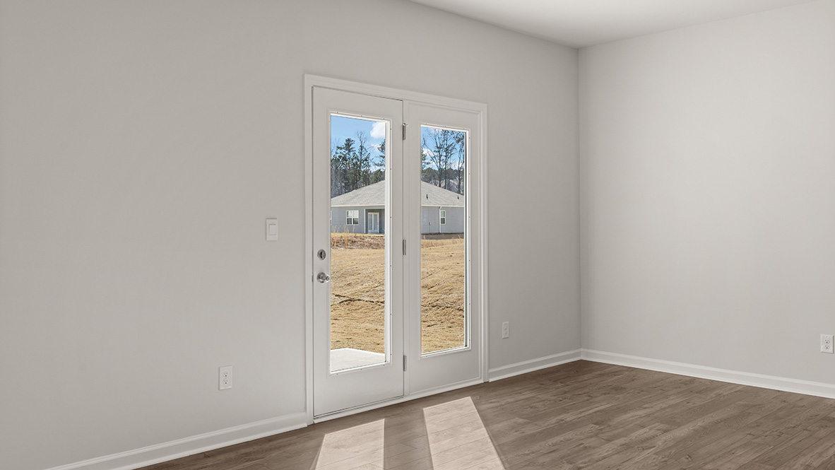A bright, empty room with light gray walls and wooden floors. A glass double door on the left shows a sunny outdoor scene with houses and trees.