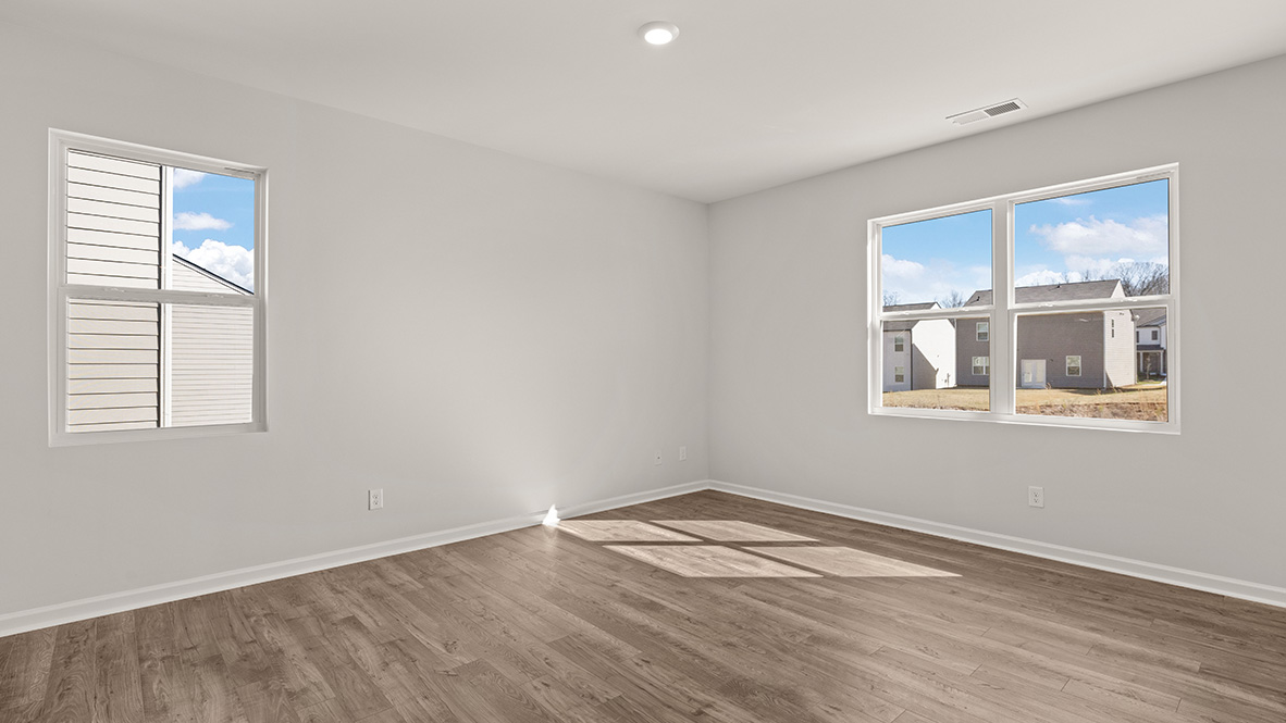 Empty room with light gray walls and hardwood flooring. Sunlight enters through two windows, casting shadows on the floor. A calm, serene space.