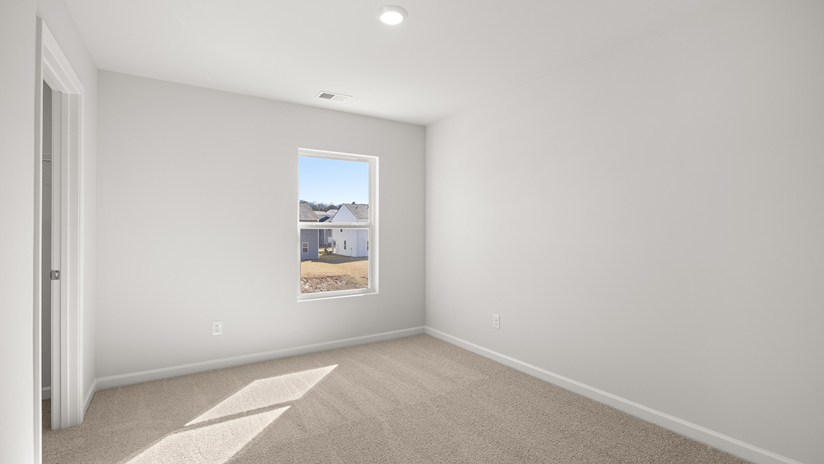 Empty room with beige carpet, white walls, and a single window showing rooftops outside. Sunlight filters in, creating a bright, neutral atmosphere.