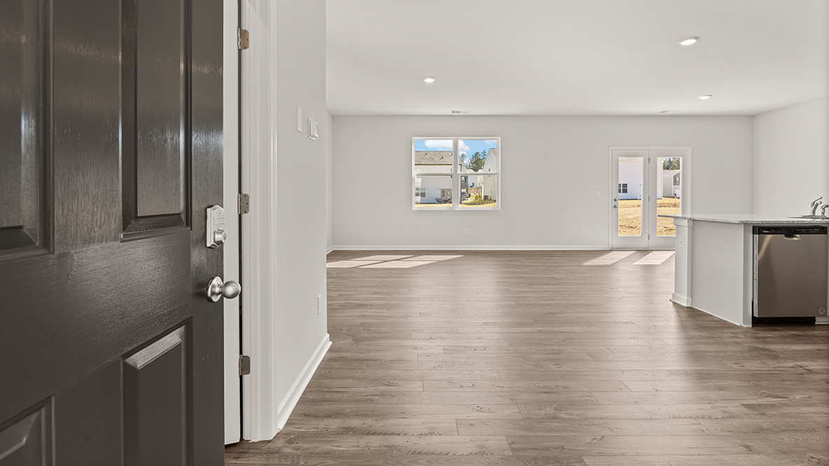 Open door leading to a bright, empty living space with light wood floors, white walls, large windows, and a kitchen area. Peaceful and welcoming atmosphere.