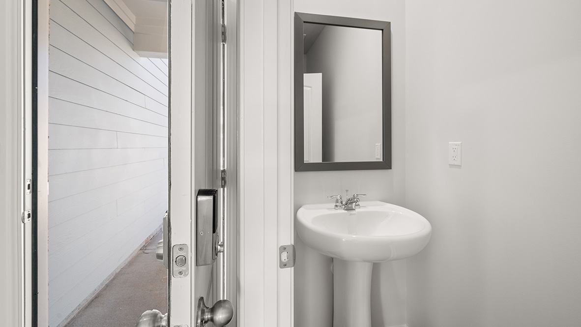 Open door reveals a small, white bathroom with a pedestal sink and a rectangular mirror. The clean, minimal design conveys simplicity and freshness.