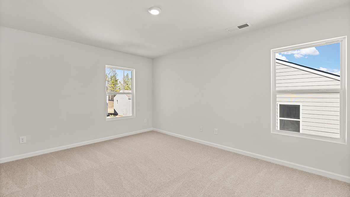 Empty room with beige carpet and white walls. Two windows show a view of a neighboring house and trees under a clear blue sky. Bright and serene.