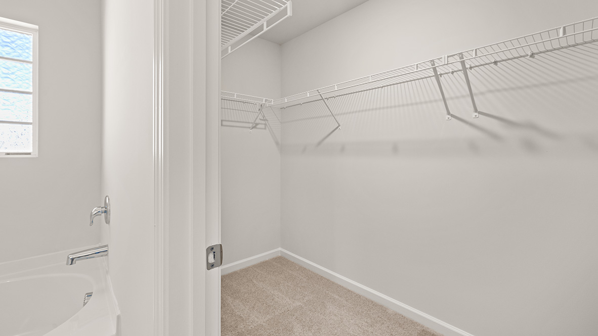 Walk-in closet with beige carpet, white walls, and two wire shelves. Sunlight filters through a frosted window, creating a bright, clean atmosphere.