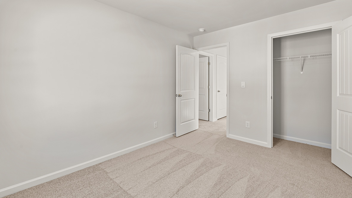 Empty room with beige carpet and white walls, featuring an open closet with a shelf and two doors ajar, creating a clean, spacious feel.