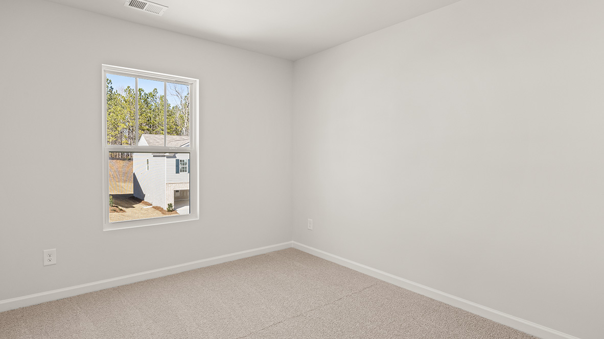 A small, empty room with beige carpet and light gray walls. A window on the left shows a house and trees outside, creating a bright and airy feel.