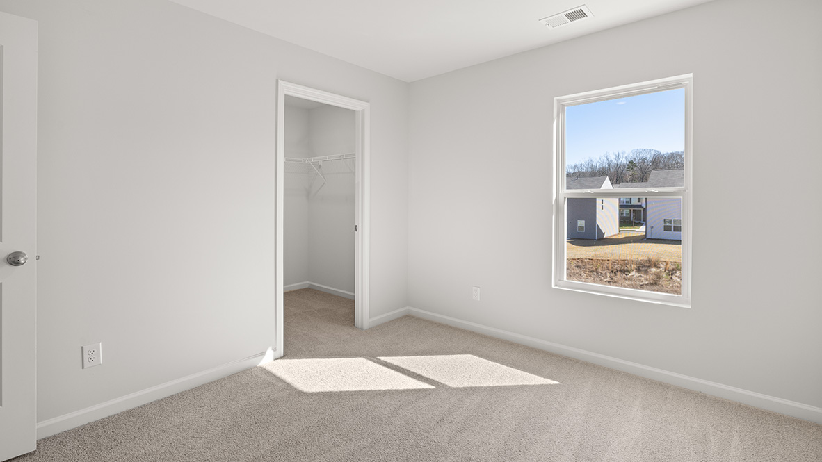 Empty room with light gray walls and carpet, featuring a window overlooking suburban houses. A small walk-in closet is visible. The room feels bright and spacious.