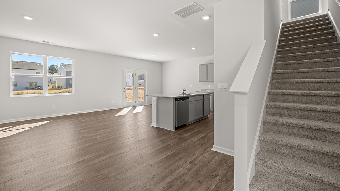 A bright, empty living area with wood floors, gray kitchen island, and stairs on the right. Large windows and glass doors let in natural light.