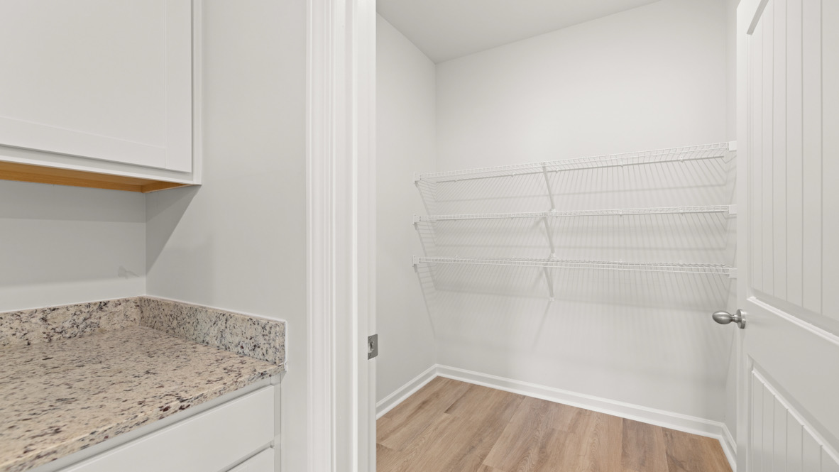 A small, empty pantry with white shelves on the right wall, wood laminate flooring, and a granite countertop on the left. The space feels clean and minimal.