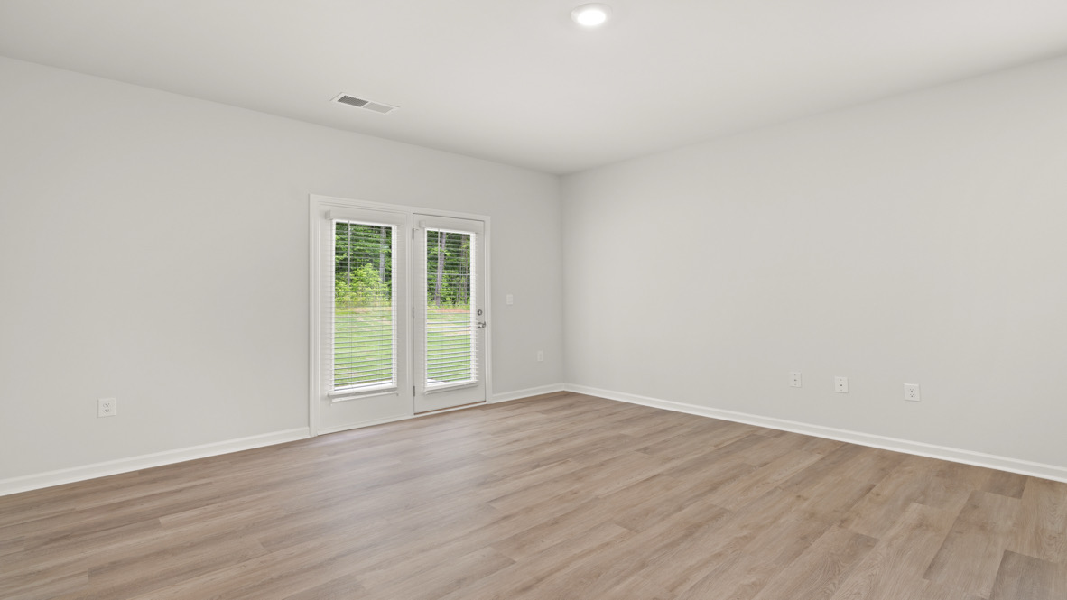 Empty room with light gray walls and wood flooring, featuring French doors with white blinds leading to a view of a green lawn and trees outside.