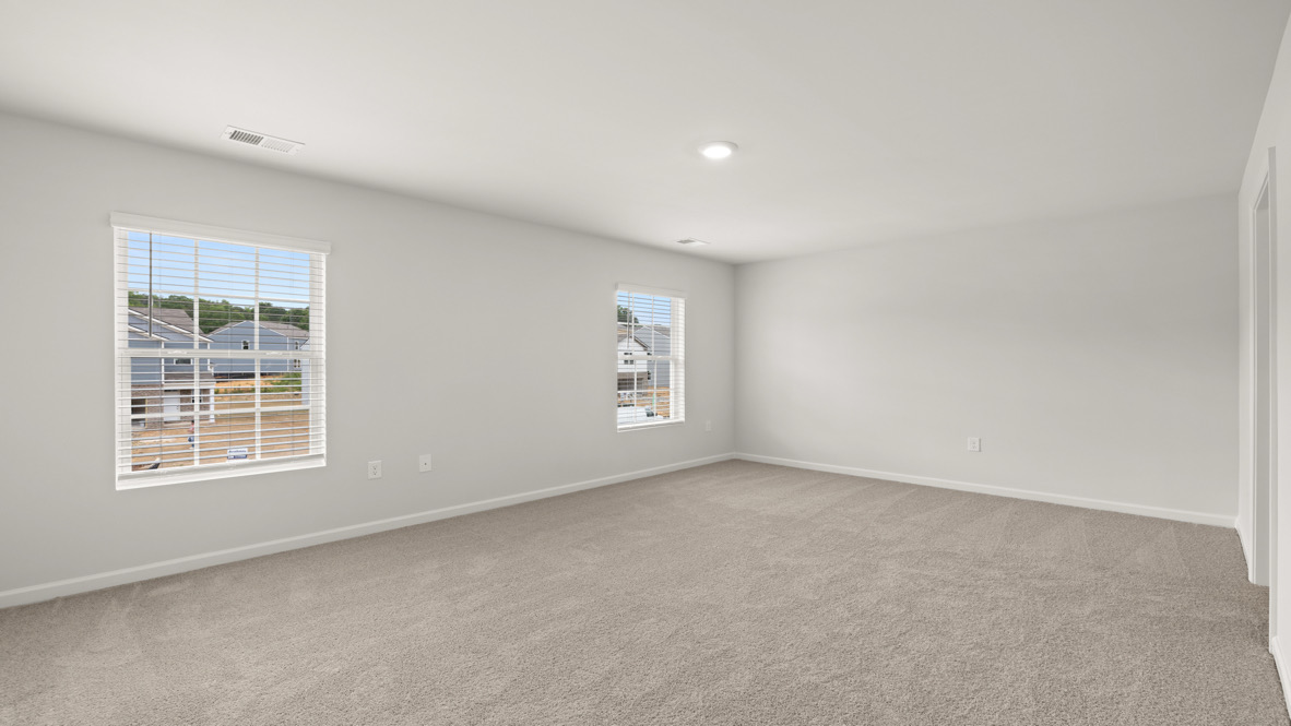 Spacious empty room with beige carpet and light gray walls. Two windows with blinds overlook suburban rooftops, providing natural light and a serene view.