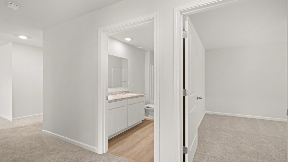 Hallway with beige carpeted floors leads to a bathroom and an empty bedroom. The bathroom has wooden floors, white cabinets, and a visible toilet. Bright lighting.