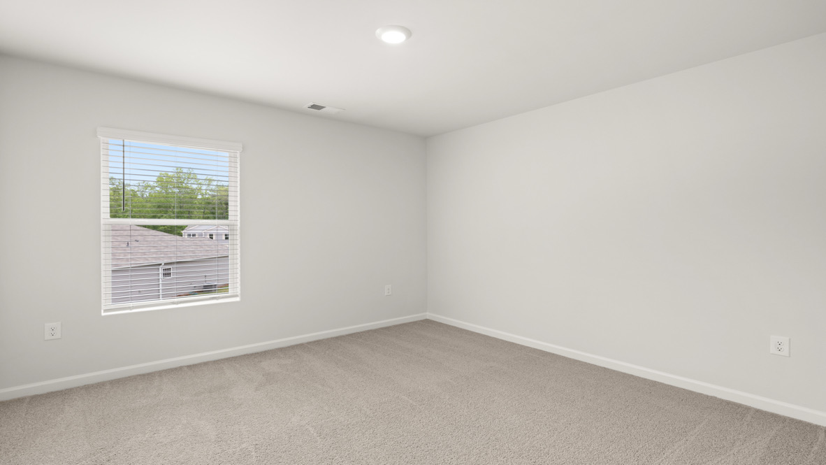 An empty, neutral-toned room with beige carpet, white walls, and a single window showing rooftops and trees outside. The space feels calm and minimalist.