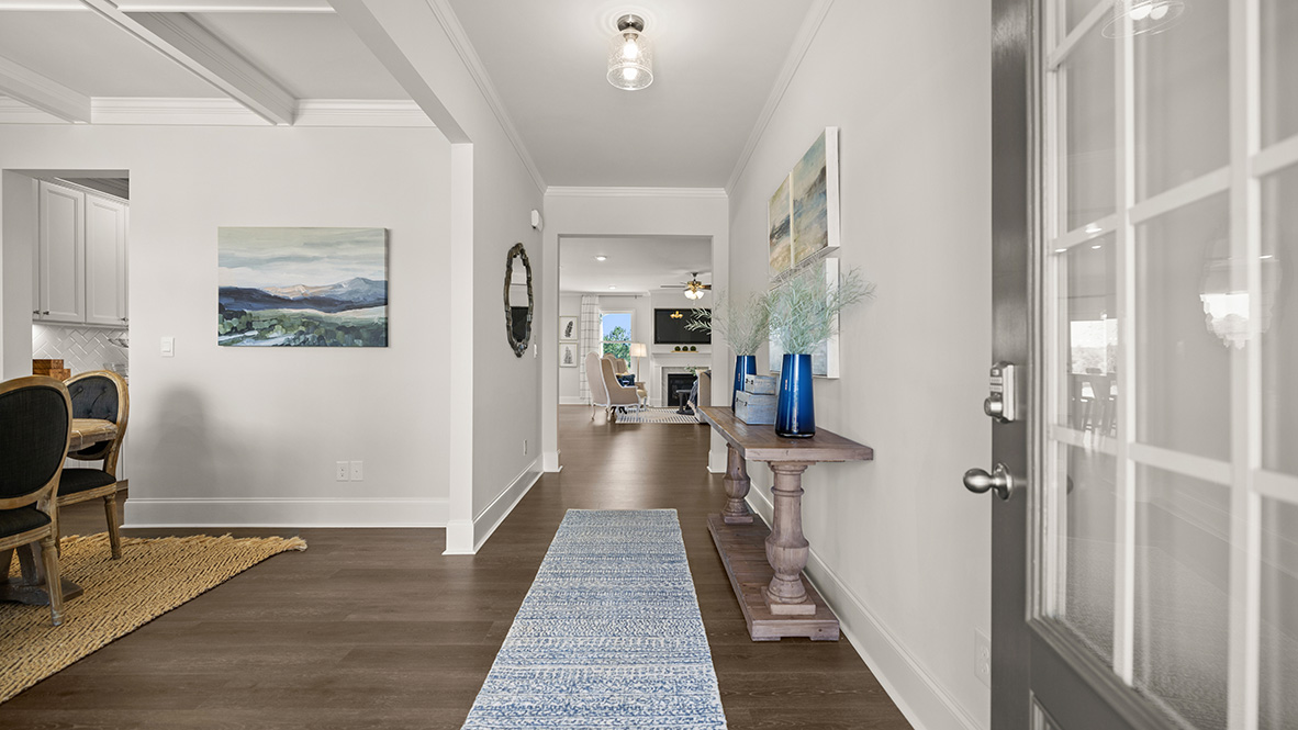 Modern home entryway with wooden flooring, white walls, and recessed lighting. Visible kitchen entrance and distant living area with a fireplace.