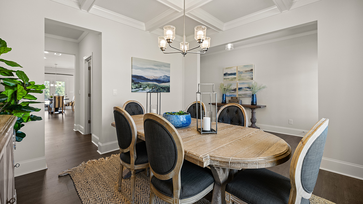 Modern dining room with white walls and coffered ceiling. A silver chandelier emits warm light over the wooden floor, creating an elegant ambiance.