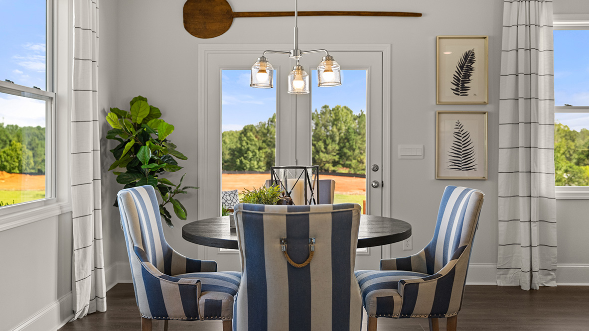 Bright, empty room with light gray walls, wooden floor, and a chandelier. Large windows and glass doors reveal an outdoor lawn. Calm and inviting atmosphere.