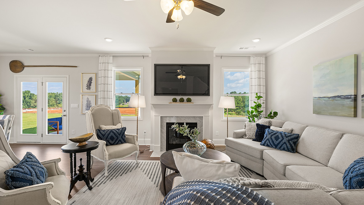 Spacious living room with hardwood floors, white walls, and large windows. Features a fireplace, ceiling fan, and modern kitchen area.