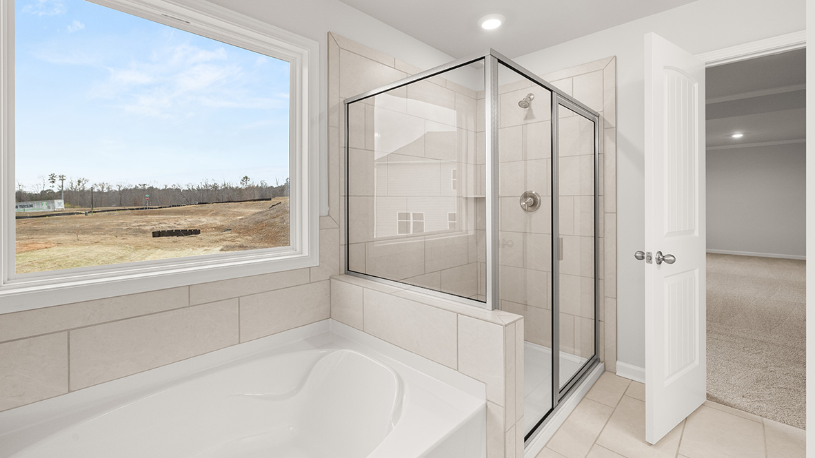 Bright bathroom with a large window showing a scenic field, sleek glass shower with beige tiles, and a white bathtub. The door leads to a carpeted room.
