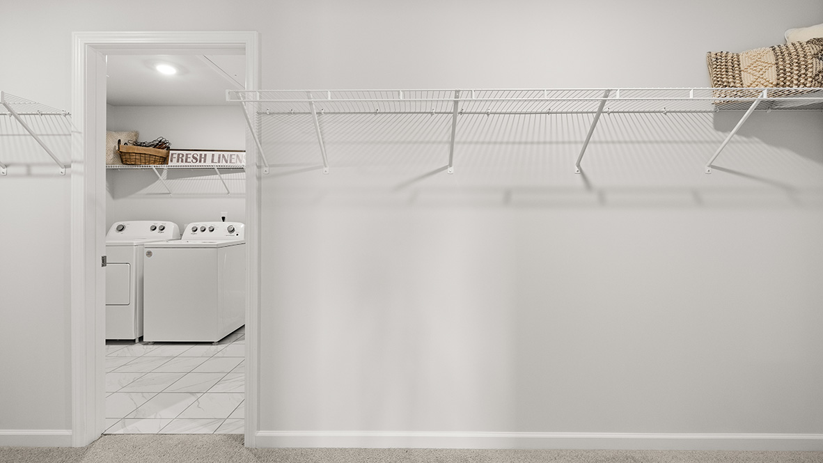 A spacious, empty walk-in closet with beige carpet, white walls, and white wire shelving on the right. Open door reveals a brightly lit room. Minimalist feel.