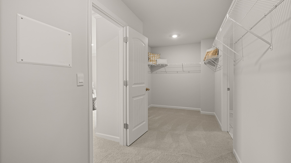 A spacious, empty walk-in closet with beige carpet, white walls, and white wire shelving on the right. Open door reveals a brightly lit room. Minimalist feel.