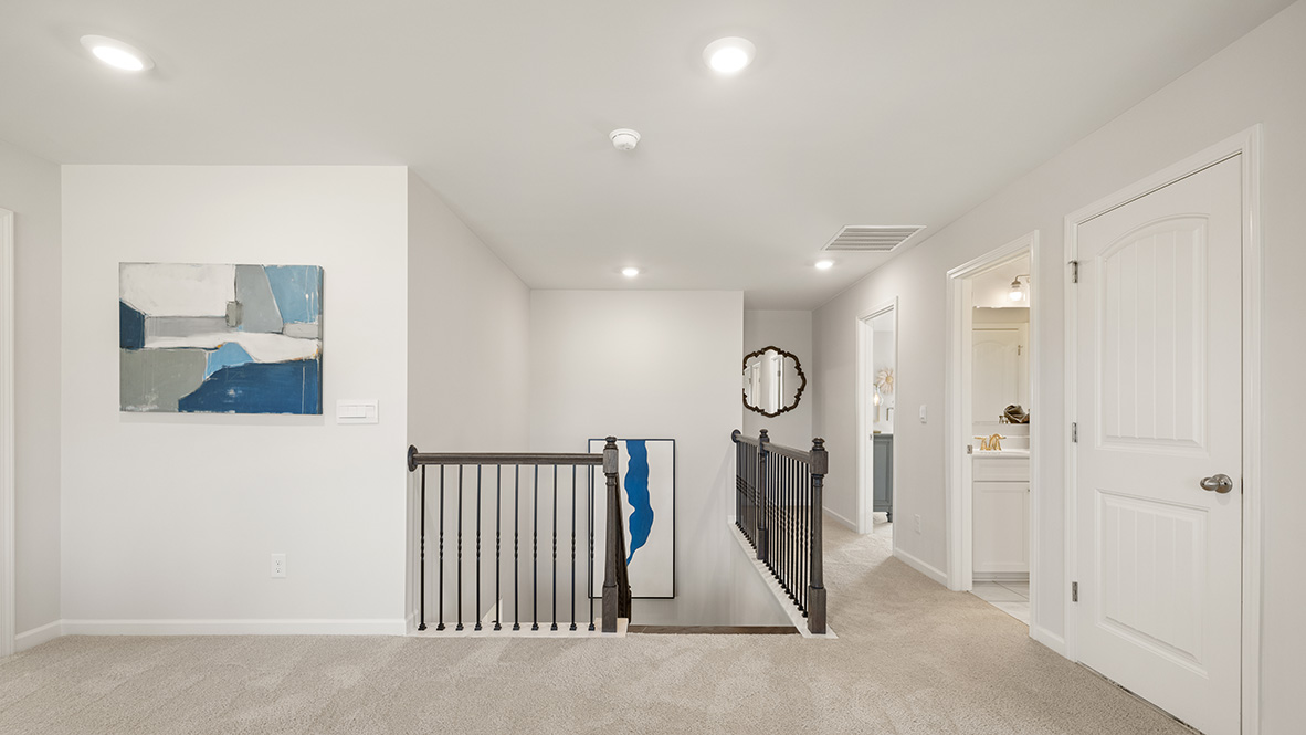 Spacious upstairs hallway with beige carpet, white walls, and several doors leading to rooms. A wooden railing borders a staircase. Bright lighting.